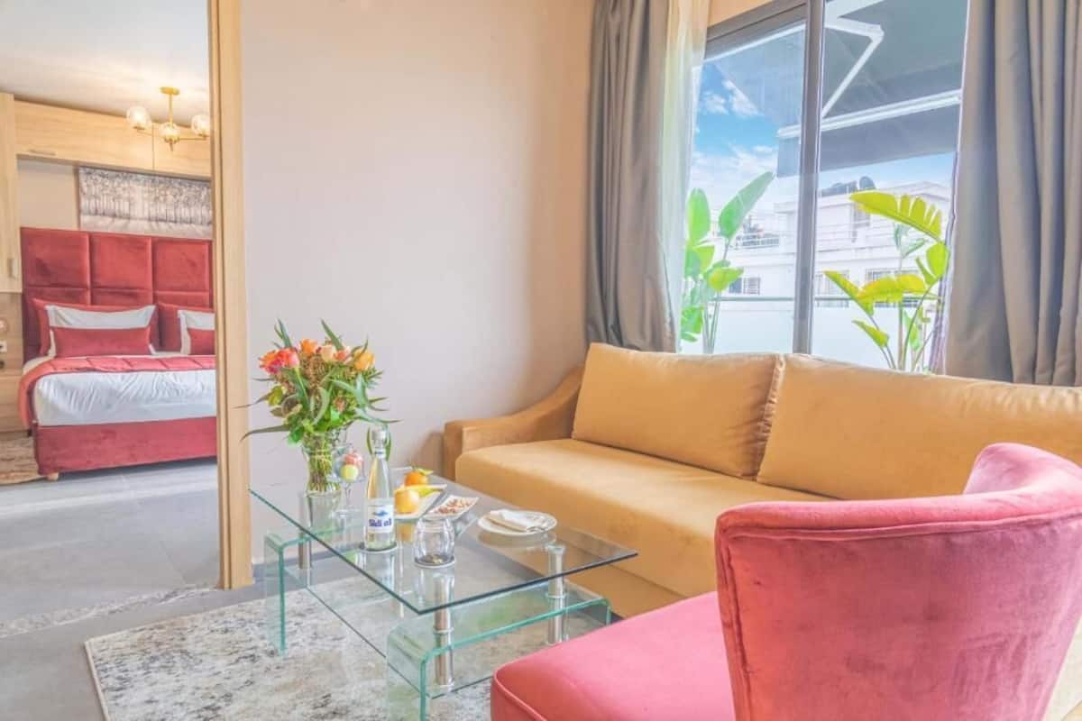 A modern hotel room with a red bed, a beige sofa, a glass coffee table, and a view of the city through large windows.