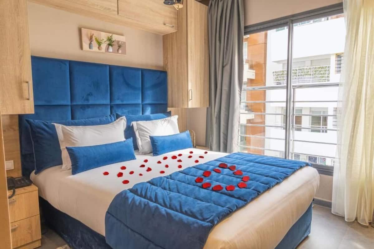 A hotel room with a large bed, a wooden headboard, and a view of the city through large windows.