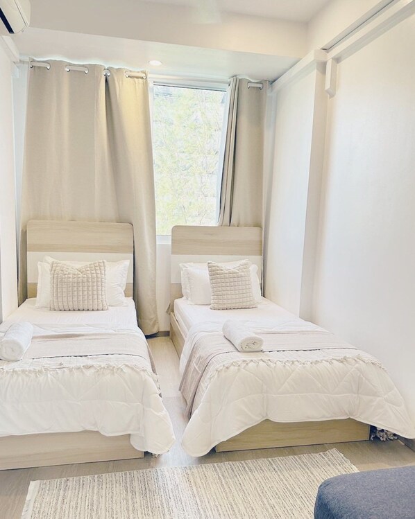 Une chambre avec deux lits, chacun doté de draps blancs et d’une tête de lit, une fenêtre avec des rideaux et un tapis de couleur claire sur le sol.