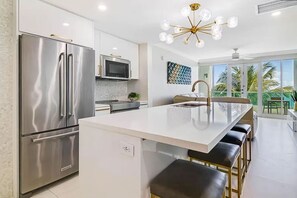 Een moderne keuken met roestvrijstalen apparatuur, een centraal eiland met barstoelen en een groot raam met uitzicht op palmbomen.