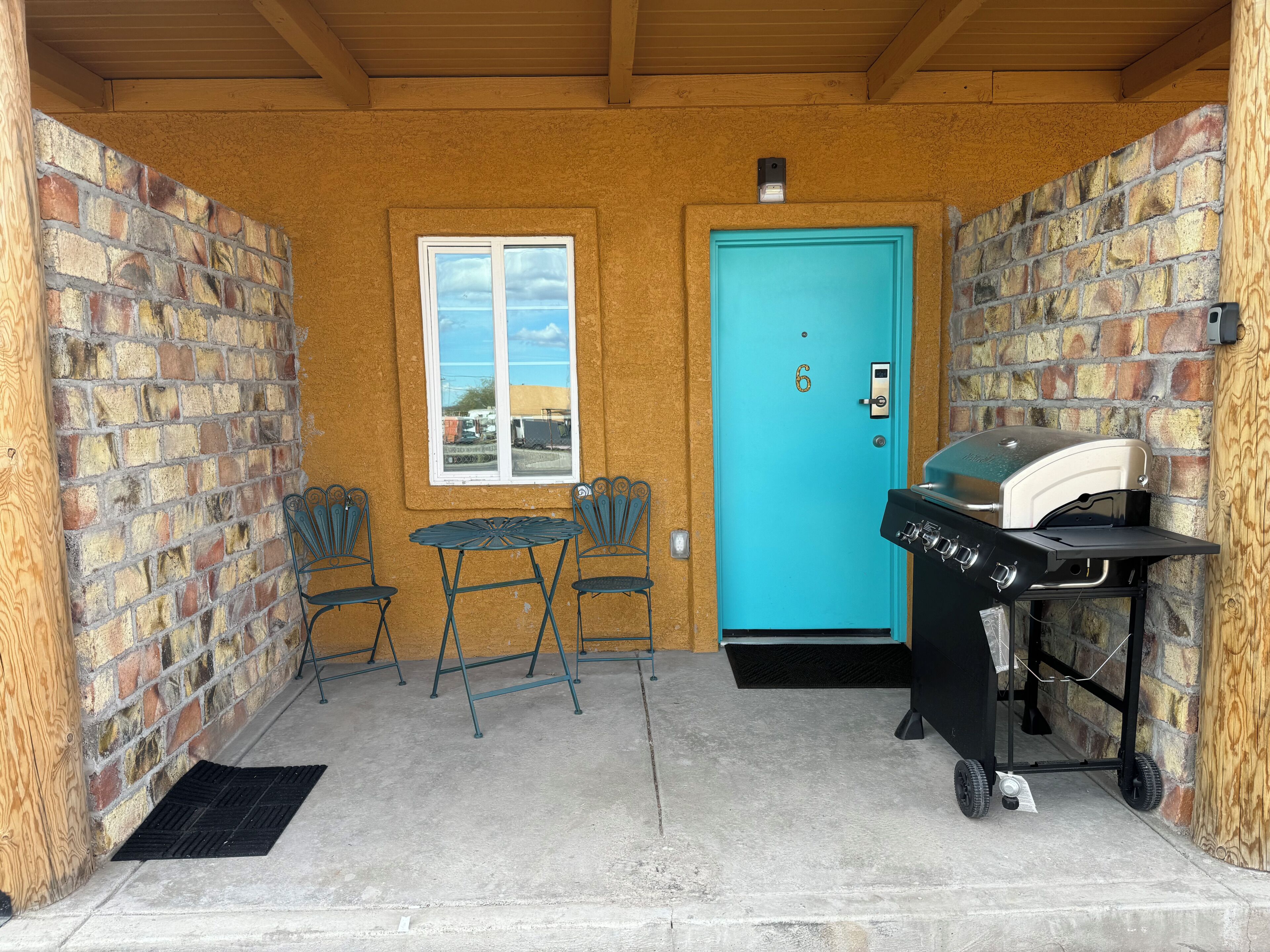 Een klein buitenplaatsje met een turquoise deur, een barbecue, twee metalen stoelen en een raam met uitzicht op een parkeerplaats.