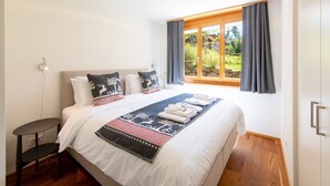 A neatly made bed with a white comforter and decorative pillows, a dark blue throw blanket, and a view of greenery through a window with curtains.