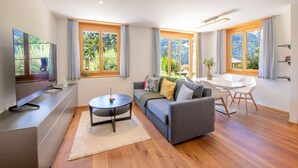 A modern living room with a sofa, TV, dining table, and large windows offering a view of greenery.