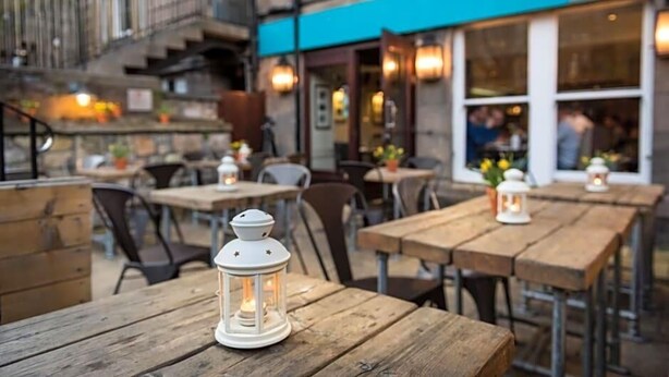 An outdoor seating area with wooden tables and white lanterns, a blue awning, and a stone building facade. - Ardgowan Hotel (St. Andrews)