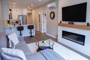 Un salon moderne avec un canapé, une table basse et une cheminée. Il y a également un coin cuisine équipé d’un réfrigérateur et d’un téléviseur fixé au mur.