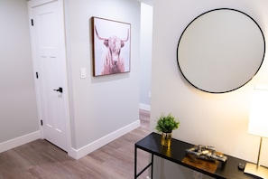A hallway with a white door, a framed artwork of a bull, a round mirror, a small table with a plant, and a lamp.