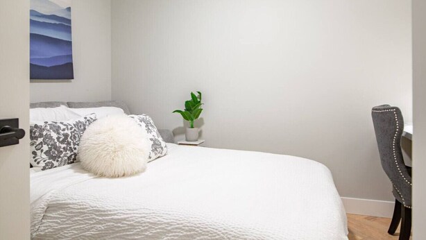 Ein Schlafzimmer mit einem Bett, einem weichen, weißen Kissen, einem grauen Polsterstuhl, einer kleinen Topfpflanze und einem an der Wand hängenden Bild.