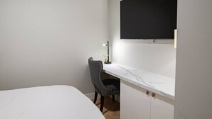 A room with a bed, a desk with a chair, a flat-screen TV mounted on the wall, and a small table with a lamp.