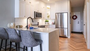 A modern kitchen with white cabinetry, a central island with bar stools, stainless steel appliances, and a herringbone wood floor.