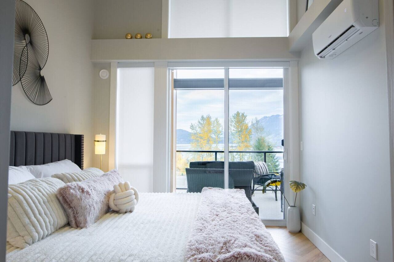 A bedroom with a large bed, a nightstand, a wall-mounted fan, and a sliding glass door leading to a balcony with outdoor furniture.