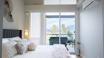 A bedroom with a large bed, a nightstand, a wall-mounted fan, and a sliding glass door leading to a balcony with outdoor furniture.