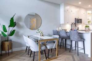 Une cuisine moderne avec des meubles blancs, un four à micro-ondes en acier inoxydable et un coin bar doté de tabourets recouverts d’un tissu gris.