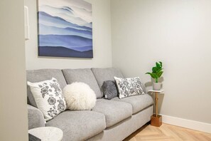 Un canapé gris agrémenté de coussins à motifs floraux et moelleux, une petite table d’appoint blanche avec une plante, et un tableau représentant un paysage de montagne accroché au mur.