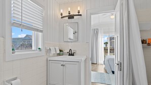 Een badkamer met een witte wastafel, een spiegel, een raam met zonwering en uitzicht op buiten.