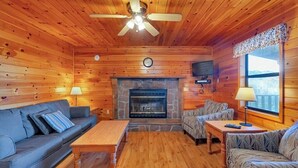 A log cabin interior with a stone fireplace, wooden walls, a ceiling fan, and a sofa. - 2 Bedroom lakeside to Dale Hollow Lake! (Byrdstown)
