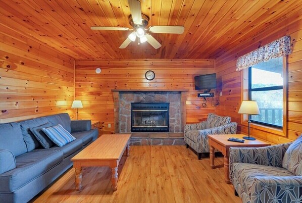 A log cabin interior with a stone fireplace, wooden walls, a ceiling fan, and a sofa.