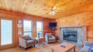 A cozy cabin interior with wooden walls, a stone fireplace, two patterned armchairs, a wooden coffee table, and a ceiling fan. - 2 Bed Chalet Lakeside with Exceptional View of Dale Hollow Lake  (Byrdstown)