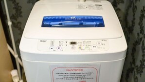 Máquina de lavar Haier branca com visor digital e painel de comando, apresentando uma etiqueta de advertência em idioma estrangeiro.