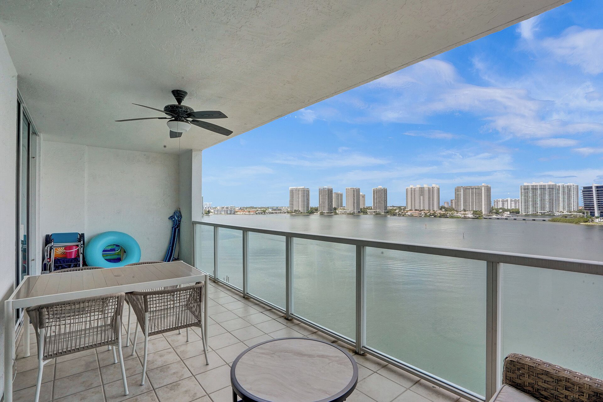 Een balkon met een tafel en stoelen, een plafondventilator en uitzicht op een stadshemelbeeld over het water.