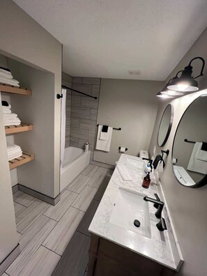Une salle de bain moderne équipée d’une baignoire, d’une douche, d’un meuble-lavabo avec un miroir et d’étagères où sont rangées les serviettes.