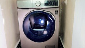 Una lavadora compacta de acero inoxidable con carga frontal en un pequeño cuarto de lavado con piso de baldosas y un cesto de basura.