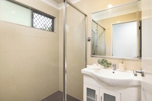 Un baño moderno con mampara de vidrio, lavabo blanco con espejo y un ventanal pequeño con celosía metálica.