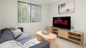 A modern living room with a grey sofa, a wooden coffee table, a TV on a wooden stand, and a window with vertical blinds. - Comfortable Rocklea 1BR | Near Shops | Parking | 173B2 (Rocklea)