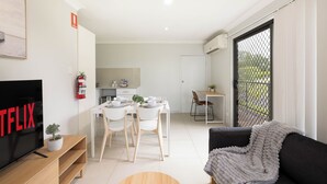 A modern living room with a flat-screen TV, a dining area with white chairs and a table, and a large window with a view of greenery. - Comfortable Rocklea 1BR | Near Shops | Parking | 173B2 (Rocklea)