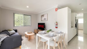 A modern living room with a sofa, a dining table set for four, a TV displaying 'NETFLIX', and a kitchen area visible through an open door. - Comfortable Rocklea 1BR | Near Shops | Parking | 173B2 (Rocklea)