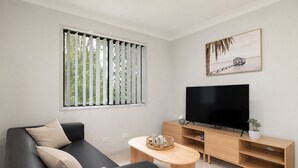 Un salon moderne avec un canapé noir, une table basse en bois, un téléviseur posé sur un meuble en bois et une fenêtre équipée de stores verticaux.