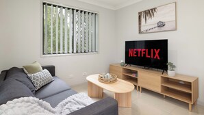 Een moderne woonkamer met een grijze bank, een houten salontafel, een televisie op een houten dressoir en een raam met verticale jaloezieën.