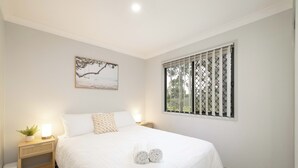Een keurig opgemaakte slaapkamer met een bed, een nachtkastje met een lamp, een kleine plant en een raam met jaloezieën.
