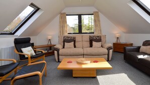 A cozy loft bedroom with a sloped ceiling, a large window with curtains, a sofa, a chair, a small table, and a lamp. - Heronlea Cottage -  Picturesque Scottish setting, 4 guests, pet friendly,  (Dunoon)