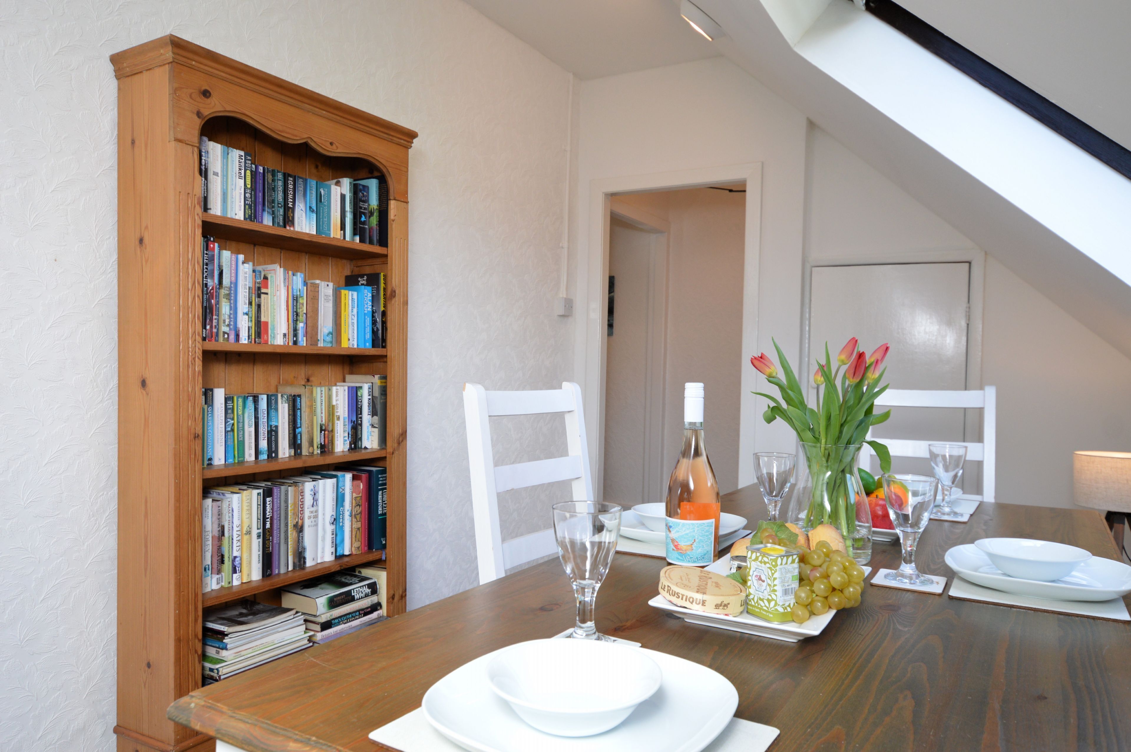 A dining table set with plates, glasses, a bottle, and a vase with flowers. A wooden bookshelf with books is to the left.