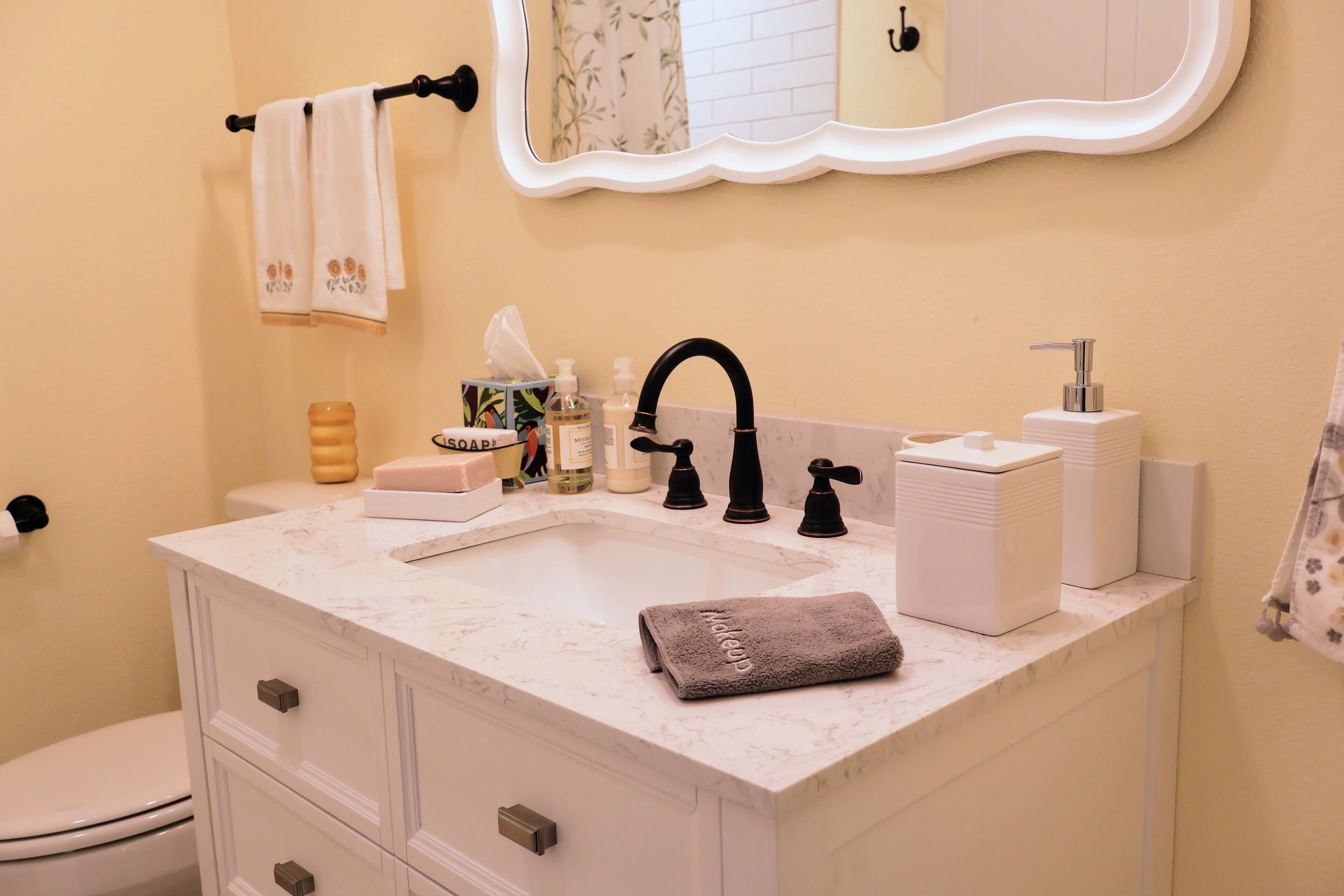 A bathroom with a white marble sink, black faucet, and a mirror above. There are toiletries on the counter and a towel hanging on the wall.