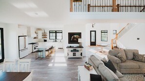 Amplia sala de estar con techo alto, cocina con armarios blancos, comedor con mesa y sillas, y una escalera que conduce a un nivel superior.