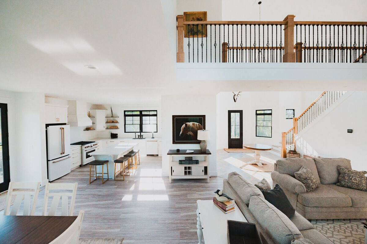 A spacious living area with a high ceiling, a kitchen with white cabinetry, a dining area with a table and chairs, and a staircase leading to an upper level.