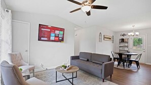 Un salon avec un canapé gris, un fauteuil beige, une table basse, un ventilateur de plafond, un téléviseur fixé au mur et un coin repas avec une table et des chaises.