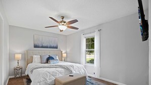 Une chambre à coucher avec un lit, un ventilateur de plafond, une fenêtre avec des rideaux, un tableau au mur et un tapis.