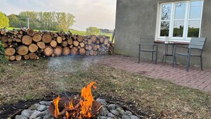 Un foyer avec des bûches qui brûlent, une pile de bûches coupées, une table avec des chaises et une fenêtre donnant sur un paysage verdoyant.