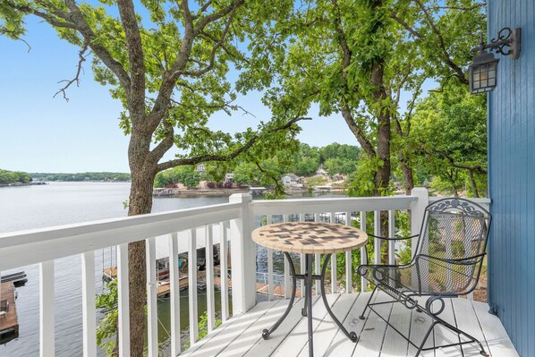 A wooden deck with a metal table and chair, overlooking a lake with boats and a dock.