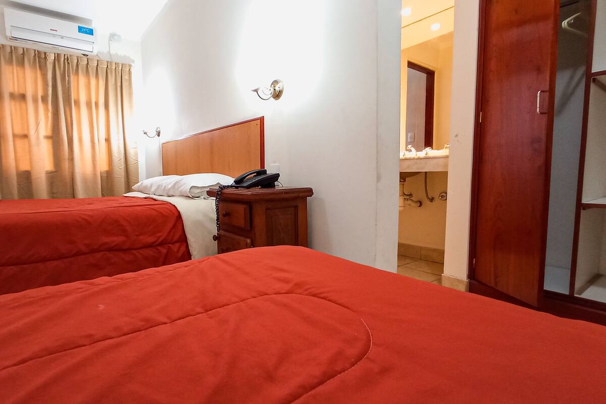 Une chambre d’hôtel avec deux lits, une table de chevet en bois avec un téléphone, une lampe fixée au mur et une salle de bain visible à travers une porte ouverte.