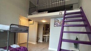 Un dormitorio con literas, una escalera morada, una lámpara en el techo y una zona de comedor al fondo.