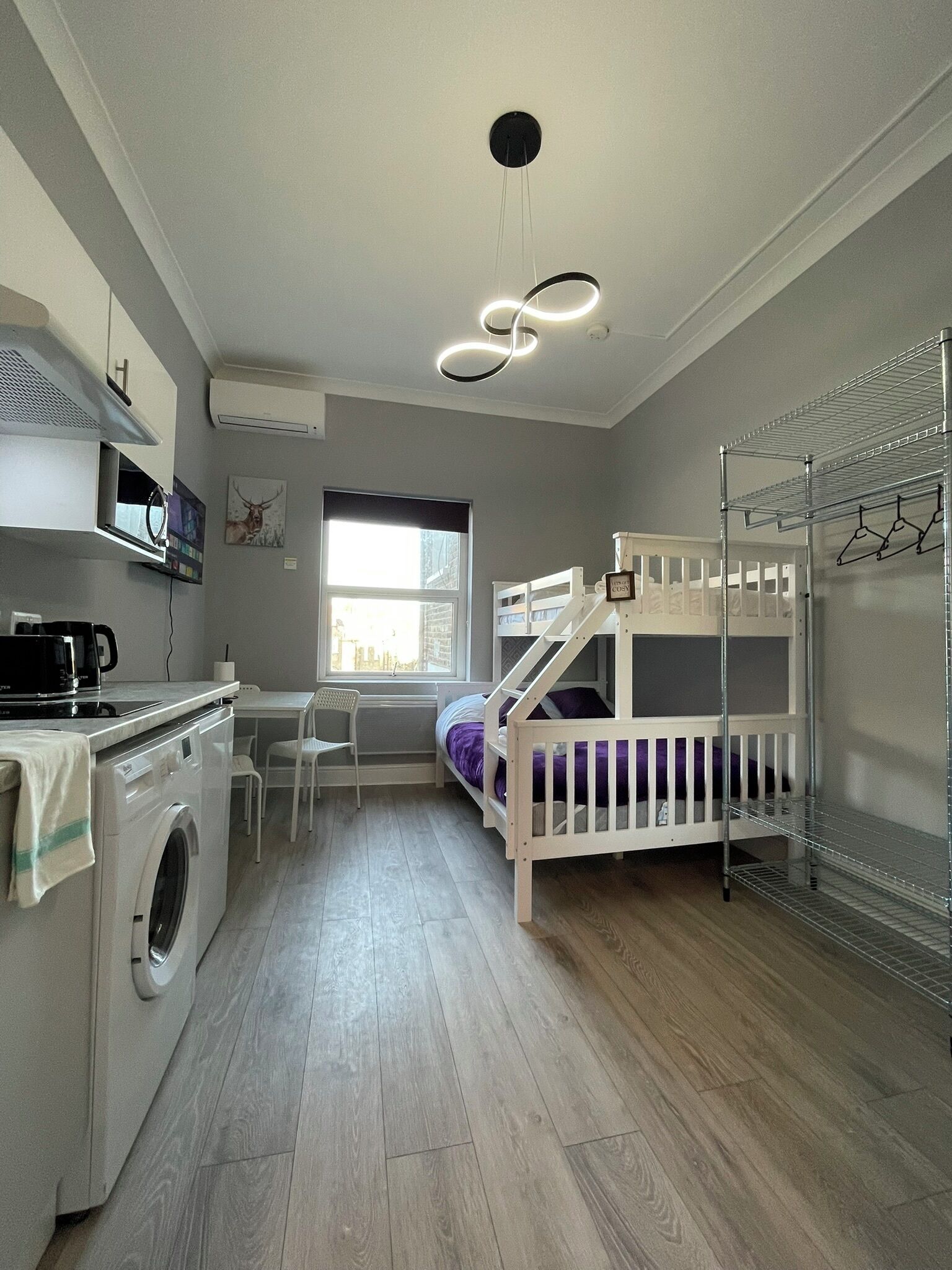 Een kleine kamer met een stapelbed, een wasmachine, een tafeltje en een raam.