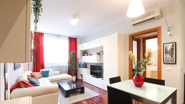 A modern living room with a sofa, coffee table, and dining table. There is a TV, a plant, and a red vase on the table.