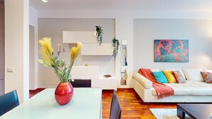 A modern living room with a white sofa, colorful cushions, a red vase with dried plants, and a painting on the wall.