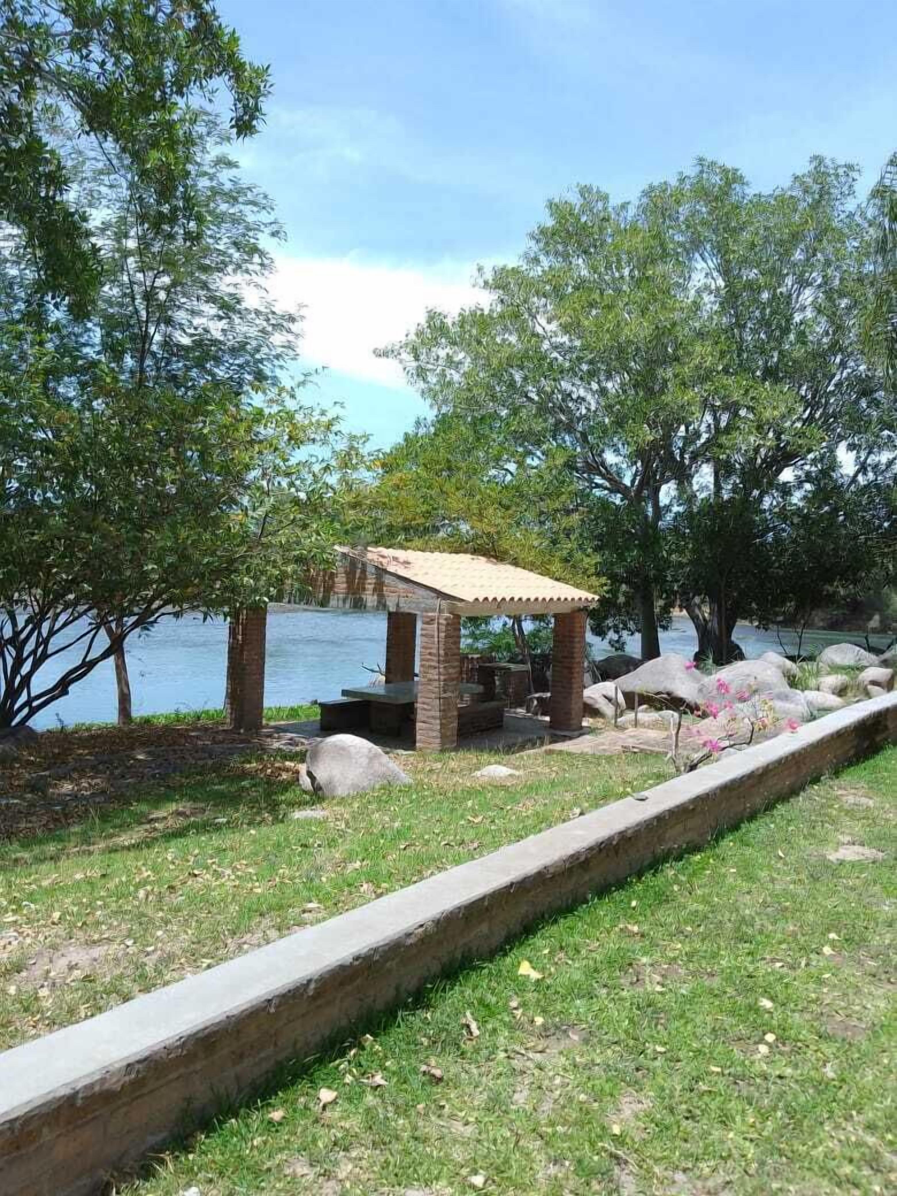 A gazebo with a tiled roof and stone pillars, surrounded by a grassy area with a stone border, near a body of water and trees.