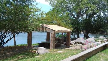 A gazebo with a tiled roof and stone pillars, surrounded by a grassy area with a stone border, near a body of water and trees.