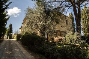 Un bâtiment rustique en pierre, auquel on accède par un chemin de terre, entouré d’arbres et d’arbustes.
