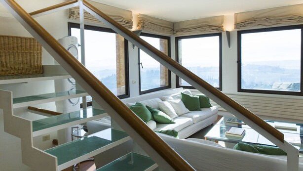 A modern interior with a staircase, a white sofa with green cushions, and large windows offering a city view. - Zafferano Rooftop Terrace Tower House (San Gimignano)